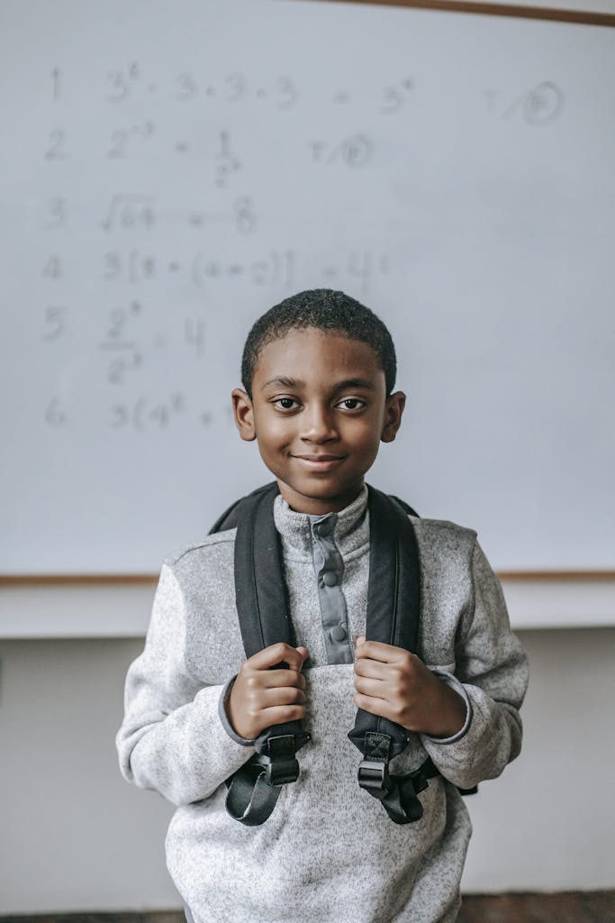 Cheerful African American boy in casual outfit with backpack standing against whiteboard in classroom and looking at camera